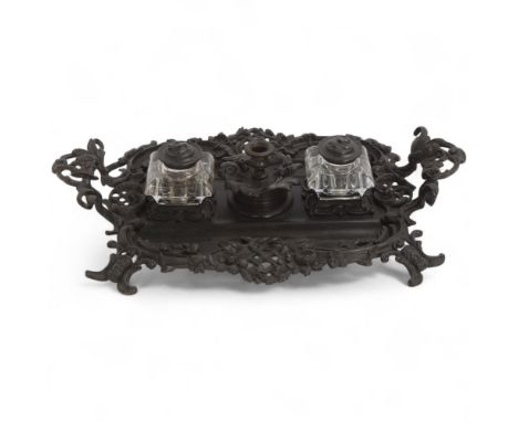 A 19th century patinated bronze desk stand, with relief cast foliate surround, original bronze lidded glass inkwells and cent