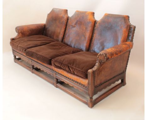 An early 20th century oak and leather three-seater sofa - the back with three shaped padded leather panels, over scroll arms 