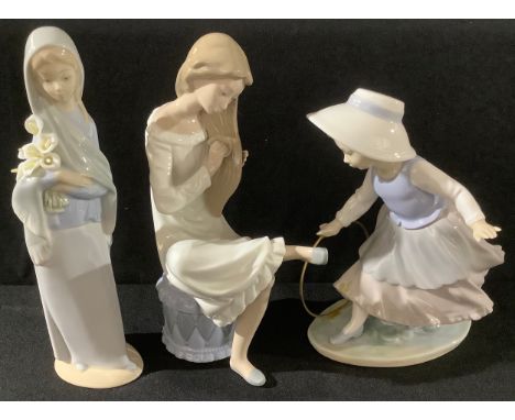 A Lladro figure, girl holding a bunch of lilies, 23cm high; a Nao by Lladro figure, young girl with hoop, 20cm high; another,