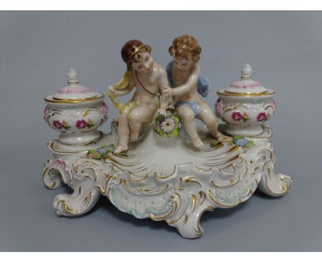 A SITZENDORF DESK TOP INKWELL, having figural seated cherubs flanked by floral decorated wells and covers with liners over sh