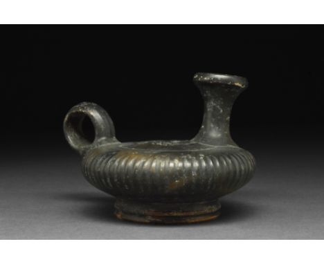 Ca. 360-320 BC.A black-glazed ceramic guttos with a discoid body, pedestal foot, loop handle, and trumpet-shaped spout; circu