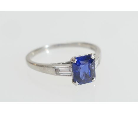 Sapphire solitaire ring, emerald cut stone of approx. 1ct, in a platinum four claw mount, flanked by shoulders inset with a b