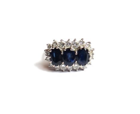 A sapphire and diamond set ring, claw set with a row of three oval cut sapphires at the centre, within a surround of circular