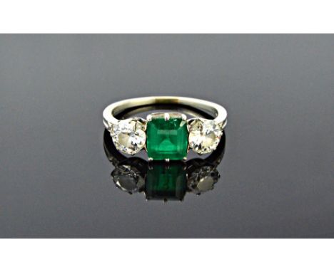 A platinum, diamond and emerald set three stone ring, claw set with the rectangular step cut emerald at the centre, between t