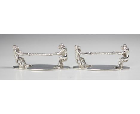 A near pair of silver plated novelty knife rests, the terminals designed as apes playing 'tug of war' upon shaped plain polis