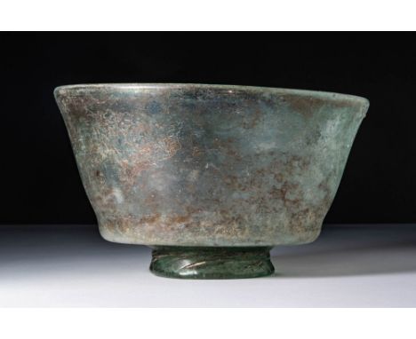 Ca. AD 1 - 200.A green glass bowl with a broad, flaring body. The lower section tapers inward and rests on a thick, separatel
