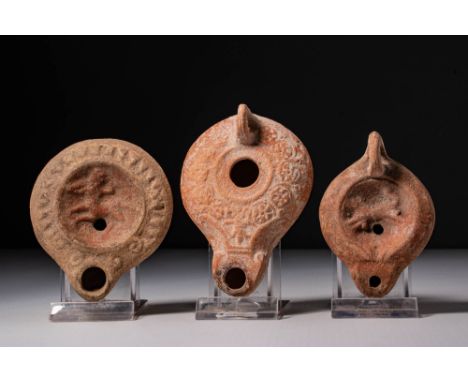 Ca. AD 1 - 200. A collection of three terracotta oil lamps. The one on the left has a flat circular body with a raised centra