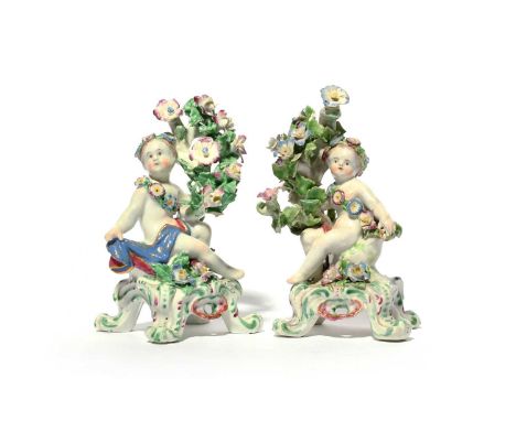 A pair of Bow figures of putti, c.1760, each seated beside flowering branches, wearing a floral sash and a diadem, raised on 