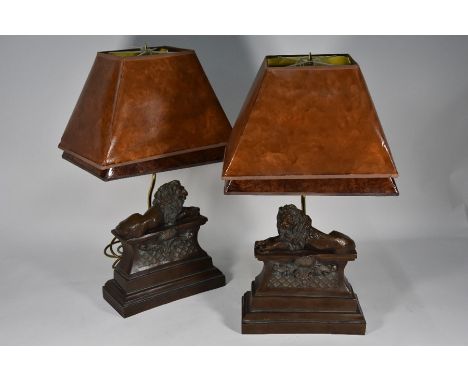 A pair of cast bronze resin table lamps as recumbent lions on plinths, by Thomas Blakemore Ltd with faux leather shades, 58 c