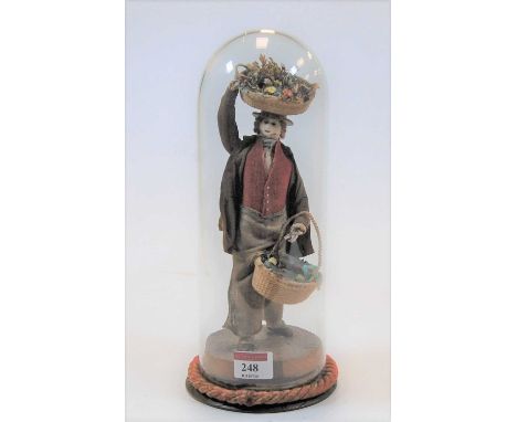 An early 20th century wire framed doll in the form of a man with a basket upon his head and another in his hand, under a glas