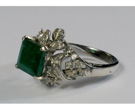 An 18ct white gold, emerald and diamond set ring, the square cut emerald mounted above diamond set heart forms, approx size M