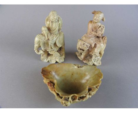 A Chinese soapstone figure holding a basket of peaches and seated upon a lion dog, Qing Dynasty, 19 th century, well carved a