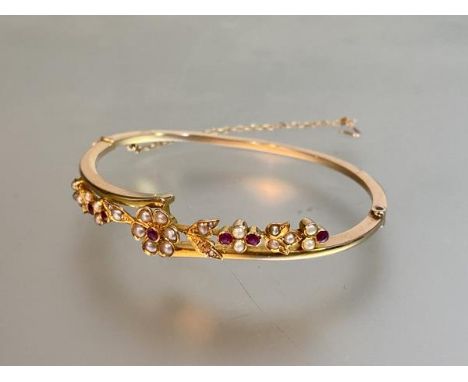 An 18ct gold bangle set with rubies and seed pearls (one lacking), early 20th century, the oval band incorporating a floral a