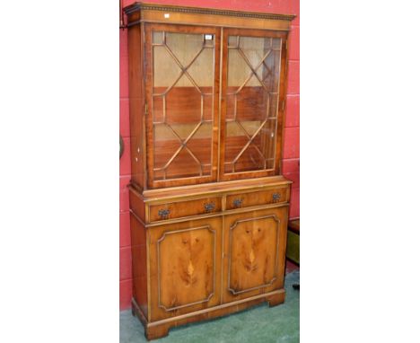 A reproduction yew veneered bookcase cabinet, dentil cornice, astragal glazed doors enclosing shelving to top, projecting bas