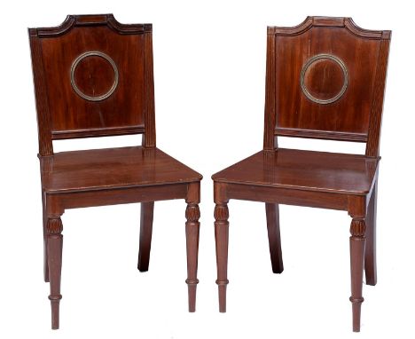 A pair of Regency brass mounted mahogany hall chairs, the panel back applied with a brass ring beneath swept tablet, having s