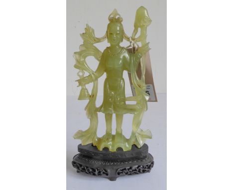 A Chinese carved light-green hardstone carving of a standing female figure, a staff in her left hand and a bell in her right,