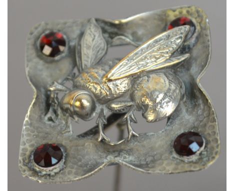 A large hammered effect metal hat pin decorated with a bee in a shaped square setting with ruby coloured stones to each corne