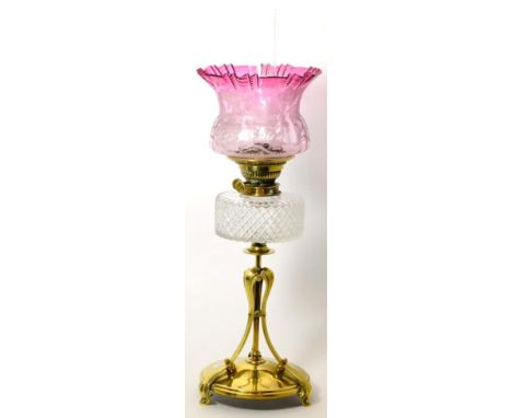 A Victorian brass oil lamp base with foliate edged cranberry glass shade, glass chimney, cut glass reservoir on triple leaf f