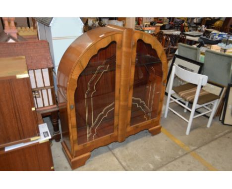 An Art Deco walnut display cabinet, of arched form, the interior glass adjustable shelves enclosed by a pair of gilt decorate