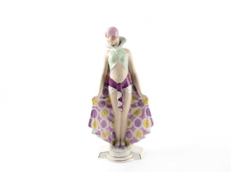 A Royal Dux Art Deco pottery figure of a female dancer with patterned high collared cape and skull cap hat. Standing on a ste