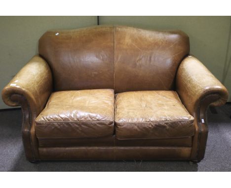 A worn brown leather sofa with arched back, scrolled stud work arm rests, raised on squat bun supports to casters, L150cm x D