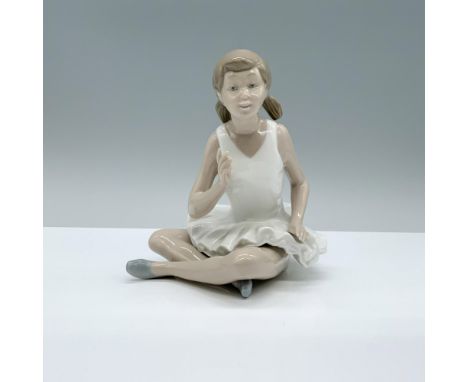 A figurine of a young girl dressed in a pristine white ballet uniform, seated with crossed legs, her hand and fingers slightl