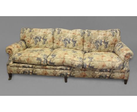 AN UPHOLSTERED THREE SEATER SOFA. in the Howard style, with scrolling arms and tapering legs on brass castors, upholstered in