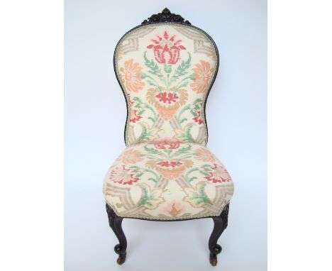 A Victorian ebonised upholstered low chair, the waisted back with foliate crest piece, on cabriole forelegs, on ceramic caste