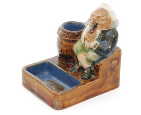 An early 20th Century Royal Doulton Lambeth vesta and ashtray formed as a seated man smoking a pipe with his arm around a bar