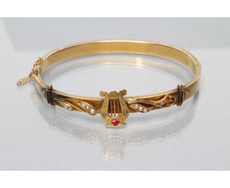 AN EDWARDIAN STIFF HINGED BANGLE, the upper section centred by an applied lyre set with a small ruby and two small rose cut d
