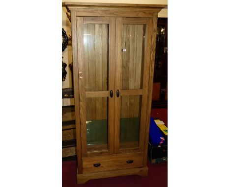 A contemporary light oak freestanding double door glazed display cabinet, having interior glass shelves over single lower dra