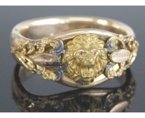 An Edwardian rolled gold hinged bangle, of good size, set with lion mask having faux ruby glass eyes, bearing armorial tablet