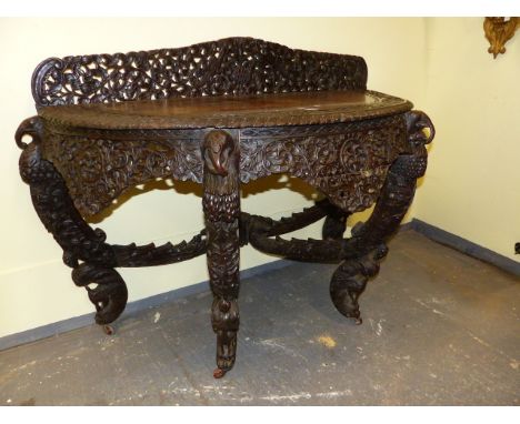 AN INDIAN HARDWOOD DEMI LUNE TABLE EXTENSIVELY CARVED WITH BIRDS, ANIMALS AND FOLIAGE, THE FOUR ELEPHANT HEADED LEGS CURVING 