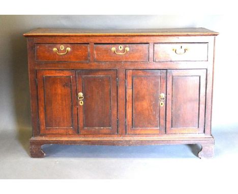 A George III Oak Dresser Base, the plank top above three drawers and four cupboard doors with brass handles, raised upon brac