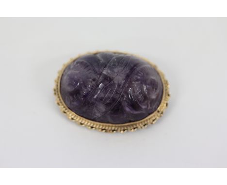 A CARVED OVAL AMETHYST BROOCH, the central stone with stylised flower head decoration, with twisted 14 carat gold frame, 3.2c