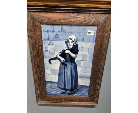 A framed Delft blue and white tile decorated with a girl holding her cat after Paul Hoecker and inscribed lower left, the fra