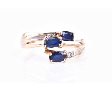 A yellow metal and triple sapphire crossover style ring, set with three mixed oval-cut sapphires and two small diamond accent