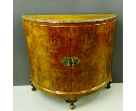 A burr walnut bow fronted side cabinet, the shaped top with carved edge over a pair of cupboard doors with brass mounts openi