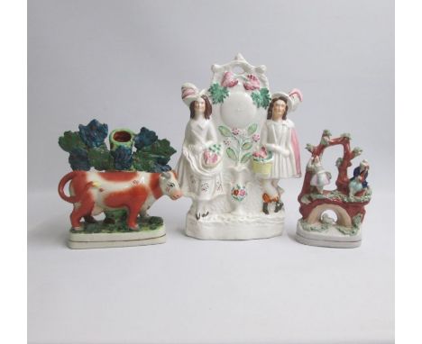 Three Staffordshire figures; a spill holder of a cow standing before bocage, a man and lady standing either side of a clock f