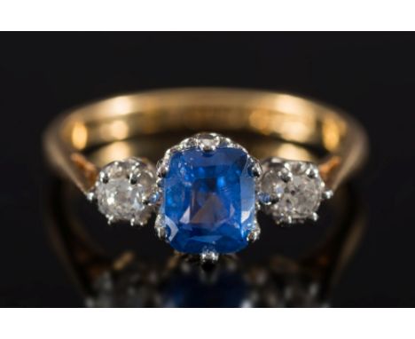 An 18ct gold, sapphire and diamond three-stone ring: with central cushion-shaped sapphire between round old brilliant-cut dia