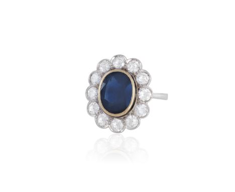 A SAPPHIRE AND DIAMOND CLUSTER RINGThe oval-shaped sapphire within a collet setting and surrounded by a border of round brill