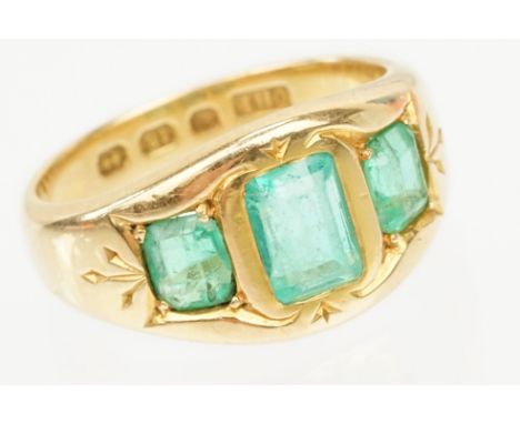 18ct gold and emerald three stone ring. The ring being bezel set with three step cut emeralds within an engraved head. Hallma