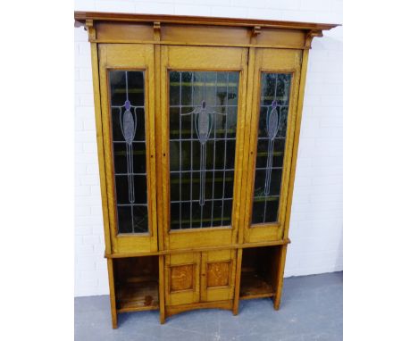 An Arts & Crafts light oak glazed bookcase, the pediment top over three lead panelled doors, above two small panelled cupboar