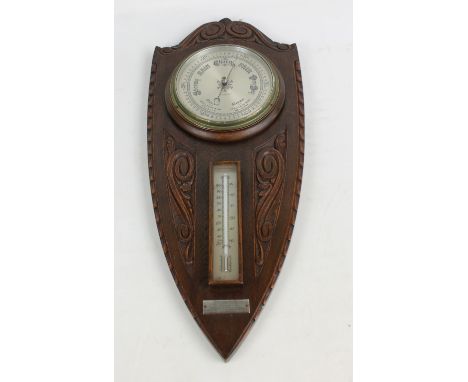 A mid-20th century carved oak barometer/thermometer in the shape of a shield with the gauge above a thermometer with presenta
