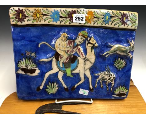 A QAJAR TILE MOULDED IN RELIEF AND PAINTED WITH A MONKEY BEHIND A MAN RIDING A CAMEL AGAINST A BLUE GROUND.   25 x 32.5cms TO