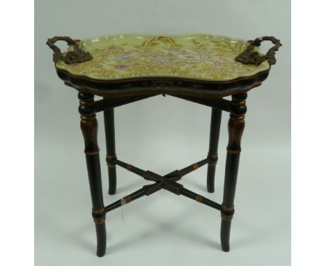 A MODERN REPRODUCTION CERAMIC TEA TRAY with cast brass handles, supported upon a Japanned and gilt decorated folding stand 
