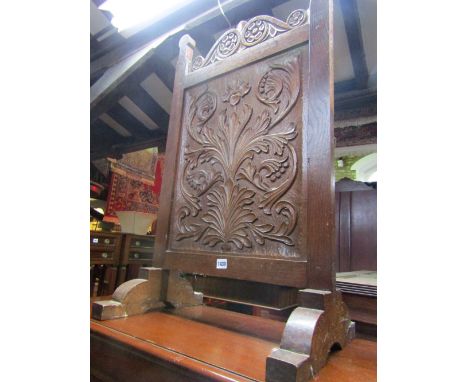 An Arts & Crafts oak firescreen with deep carved foliate panel and cresting rail raised on stepped sledge supports