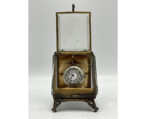 A small travel watch contained in an elaborate silver metal case with bevelled glass sides, carrying handle and small feet. T