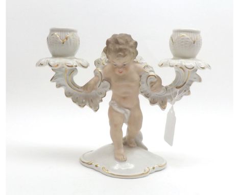 A German Porcelain Two Branch Candlestick, the central support modelled as a cupid, flesh colour and gilded detail throughout