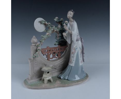 This impressive glossy figurine depicts a woman dressed in a kimono, standing in a garden and holding up a flower beneath a f